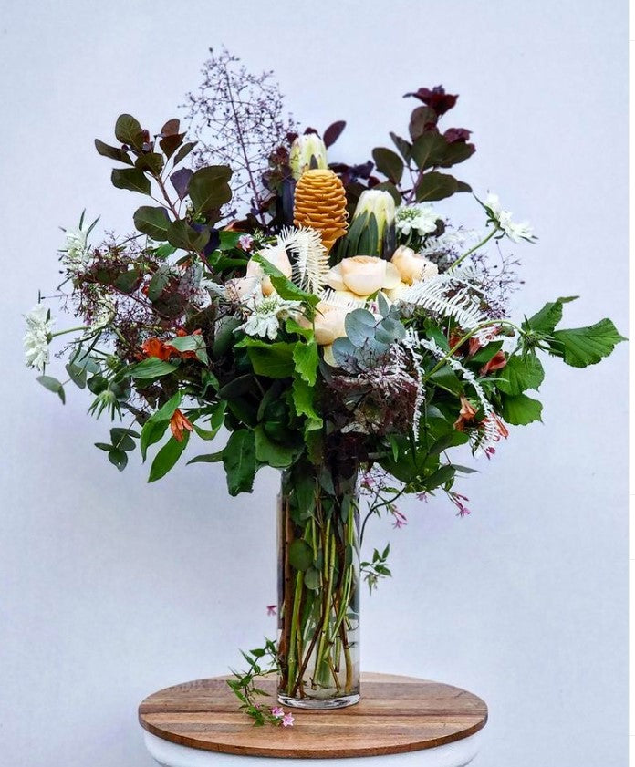 Page Elliott Floral subscription in "Yowza": Fresh flower bouquet with ginger, fragrant peach garden roses, white preserved ferns, smokebush, orange peruvian lilies, jasmine vine, white mink protea and eucalyptus. The vase sits on a wood tabletop.